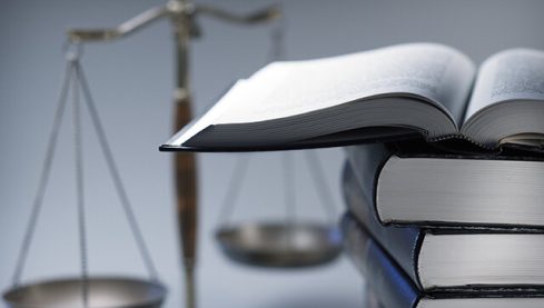 A stack of law books stands in front of a justice scale that is slightly out of focus.  On top of the stack is an open law book.