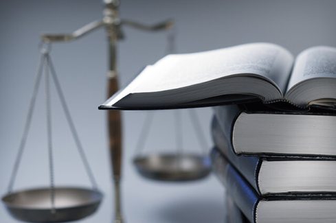 A stack of law books stands in front of a justice scale that is slightly out of focus.  On top of the stack is an open law book.